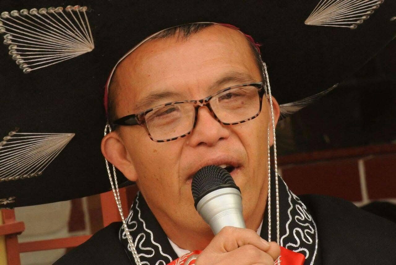 A man with glasses and a striped scarf holding a microphone while speaking